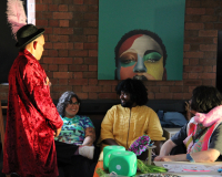 A performer in a red velvet coat and feathered hat chats with seated guests at a colorful art table beneath a bold painted portrait.