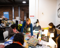 Diverse group creates art around a craft table in a bright studio, with paints, collage materials, colourful projects, and a mural behind them.