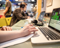 An office work place scene. In the foreground a hand rests on an open laptop holding a pen and paper. In the background out of focus another person sits at a desk wearing headphones 