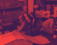 In a red-tinted workshop, several people crouch around large sheets on the floor, focused on a project beside equipment and circular artwork.