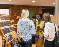 A Black woman with a soft smile and french plaits smiles softly in a conversation with two white women with blonde hair who have their backs to the viewer.