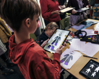 A child in a red hoodie uses a tablet to design clothing at a busy fashion workshop, surrounded by sketches, fabric swatches, and other participants.
