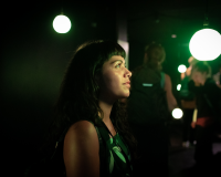 An image of a person with long black hair and a short fringe looking into the distance in a dark room, they are lit up in green neon light, and there are bright light bulbs scattered around in the background 