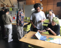 Image of seven people in the Pervasive Media Studio events space. A group of two are facing the camera and intently looking down at a book. Another group of four people, with backs to the camera, are also looking down at another copy of the same book. Another person standing on their own in the background looks into the distance.