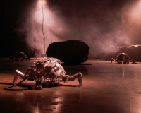 A small robotic tortoise in a dimly light room. In the background are large black boulders, smoke and another robotic tortoise. 