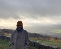 Geraint is wearing a yellow patterned beanie and grey coat. He stands against a dramatic, cloudy sky overlooking green hills and valleys in the distance.