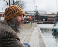 A white man with a beard and a yellow wool hat looks out over the harbourside in Bristol