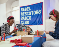 A young Black women sits smiling with red headphones on and a banner above her reads Rebel Resistors Radio Club. 