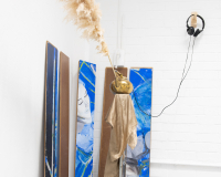 Art installation with pampas grass in a gold vase on a stand, draped with metallic fabric. Three abstract panels in blue and brown lean against a white wall. Headphones hang nearby.