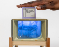A hand rests on a small screen displaying text. Below, an old TV sits on a wooden stand, showing an abstract blue image. The scene feels nostalgic and artistic.