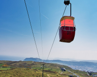 A coral coloured barrel like character sits in a cable car with a beautiful landscape behind it in Llandudno.