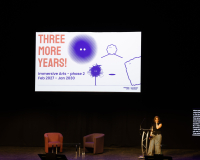 Speaker stands on a dimly lit stage beside a slide reading “THREE MORE YEARS!” with colorful drawings, creating an optimistic, creative tone.
