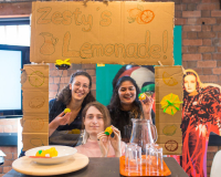 Three people are excitedly in the middle of a frame reading, 'Zesty's Lemonade' all holding lemons.