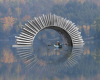 The scene is slightly misty, a man sits in a blue boat with oars on open water inside a sculpture. There are autumn trees in the background.