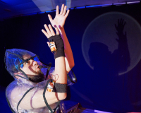 A young white man posed with his arms in the air and is wearing a see through raincoat with batteries attached to him and black fingerless gloves
