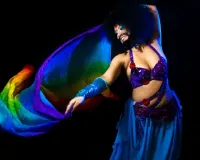 A photo of Shrouk dancing with a large piece of material with rainbow flag pattern. Shrouk is smiling, they have big black hair, bright blue eye make up and have moustache and a beard. They are wearing a belly dancer outfit; a jewel encrusted purple bra like top and a flowing blue skirt with a jewelled belt.