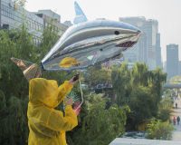 A person wearing a yellow raincoat with the hood up holding a silver shark balloon. The person is standing in front of green trees with many tall buildings in the background. To the right of the image is a path going in the city, it has a number of people walking.  