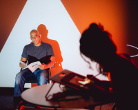 A man is reading from a book sitting in front of a wall where live illustrations are projected. The live illustrator is out of focus in the foreground.