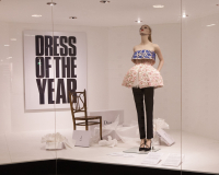 Mannequin dressed in embroidered top and short balloon skirt, propped up in window display with a poster that reads 'Dress of the Year'