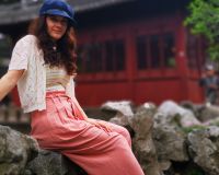 profile picture of Ellie Chadwick: woman in early 30s, sitting on a rock in front of a pagoda, with long dark hair, a blue hat, yellow striped top and pink trousers