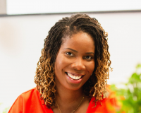 Joyann is smiling looking directly to the camera, she has should length locs and is wearing a orange dress