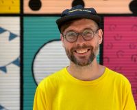 A friendly looking white man smiles in front of a colourful background. He has a brown/grey beard and glasses