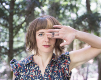 A close-up of artist Sylvia Rimat in a forest, with the palm of one of her hands over one of her eyes, revealing a tattoe-like drawing of what could be a tree or a neuron.