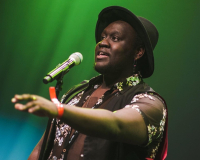 Imagine of a Black man on stage performing. The background has a green light and he is wearing a black shirt with a wide-brim hat speaking into a microphone