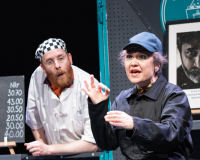 A colour photograph of a white man in his 30s with a short ginger beard and moustache wearing a black and white checkered chef's cap, and chef's whites. He's leaning over a large grey panel that serves as a serving hatch in a cafe. In front of the serving hatch is a white woman in her 40s wearing a black cap and a black boilersuit. She's miming holding a cup and the other hand signals the OK sign for divers, fingers forming a loop with the little fingers sticking up. 