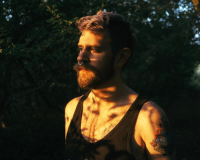 Graham is a person with a beard and a mohawk. He is stood in a dimly lit forest wearing a black vest.