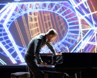 Larkhall reaches inside a piano while colourful visuals are projected behind him