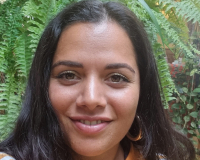 A brown woman with long dark hair and big smile. Roseanna wears a bright orange jumper and is standing in front of a lush green plant.