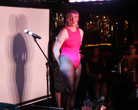 Raina is a white nonbinary person with short pink hair.  They are standing on stage next to a mic and projection screen.  She is wearing an 80's style pink leotard, black tights, and had pink hearts drawn on her cheeks.  They are looking slightly surprised, to the left of the audience.