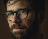 Portrait of Ben Samuels, a person with a beard and glasses, set against a dark backdrop.