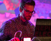 Joseph Wilk portrait, a man with dark hair and glasses sitting in front of a laptop against a pink and purple oil projector image