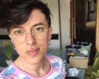 a white trans masc person with dark hair smiling slightly at the camera. they are wearing a cap, gold glasses and a t shirt striped with the colours of the trans flag. the background is the studio meeting space