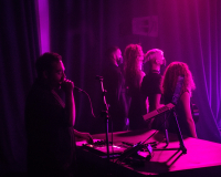 Artist Nik Rawlings, dressed in black, sings into a microphone behind a group of performers who are looking away from them, while they're all washed in pink light.