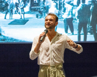 White male in a white lace shirt presenting in front of a cinema screen.