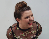 Liz, a white woman with brown hair tied in a bun is looking to her left and laughing at something off-camera, she wearing a black and embroidered flowery top and her staple red lipstick!