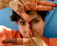 Leila against a bright blue door, her face in the centre of the image, wearing a bright orange shirt, gold hoop earrings, and black kohl eyeliner. She has short dark hair, and is looking directly at the camera, holding up both of her hands in front of her face - one with her palm-side facing the camera and one with her front-side. Her thumbs are touching at the tips of her fingertips in the centre of the image, and her hands are covered in intricate deep orange henna designs that extend onto her wrists