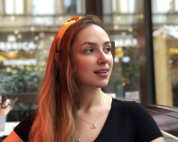 A headshot of Sasha looking past the camera. They have long light brown hair and are wearing a black T-shirt with a yellow headband.