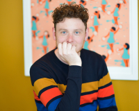 Jon is a white man with brown, curly hair. He is smiling, with his hand partially covering his mouth. He is wearing a dark jumper with three colourful stripes. He is sat behind a table looking into the camera, with colourful artwork on the wall behind him.