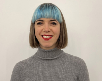 Portrait of Gabby Shiner-Hill, a smiling woman with a straight bob, blue fringe and a grey turtleneck top.