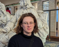 A photo of Eli, a white nonbinary person in their late 20s. They have chin length brown hair and wear round glasses. They wear a chunky black roll neck jumper and a black crossbody bag. They stand in front of a row of marble statues, on display in La Piscine Museum Roubaix.