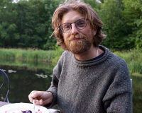 A shaggy looking thirty year old eating pie in front of a lake
