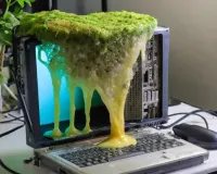 An old laptop computer sits on a desk. A green, foamy mould seeps down over the screen of the laptop.