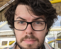 Picture of Adam Clewes-Boyne, a white man with dark brown hair, black glasses, a moustache, and beard