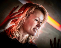 An image of artist Dr. Tine Bech delivering a talk. She is a blonde woman, with both hands raised mid speech, on either side of her head. 