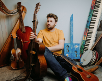 Greg wearing a yellow t-shirt sitting in amongst several musical instruments including a harp, piano, guitar and banjo
