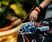 A photo of a hand wearing a rainbow wristband touching a key on a synthesizer surrounded by audio cables and guitar pedals. in the foreground there is an out of focus plant in a giant pot.